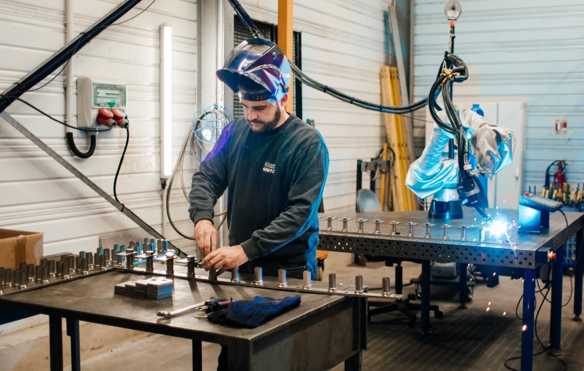 Cobot en train de souder une série de pièces pendant que le soudeur prépare le montage de la série suivante